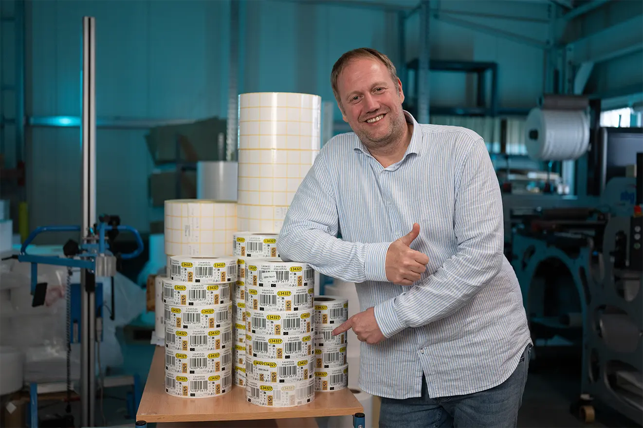 Geschäftsführer zeigt lächelnd auf mehrere gestapelte Etikettenrollen in einer Produktionshalle.
